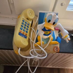 Kalle Anka fast telefon - Rolig fast telefon med Kalle Anka-design, perfekt för barnrummet eller samlare. Telefonen har knappsats och sladd, och motivet visar Kalle Anka på stranden. Mindre slitage syns på plasten, men i gott skick för sin ålder.