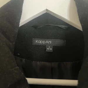 Svart kappa med huva från KappAhl - Svart kappa från KappAhl i ullblandning med huva, stora knappar och snygga detaljer vid ärmslut och axlar. Klassisk krage, dragsko vid huvan och två fickor framtill. Perfekt för kyliga dagar och riktigt stilren look.