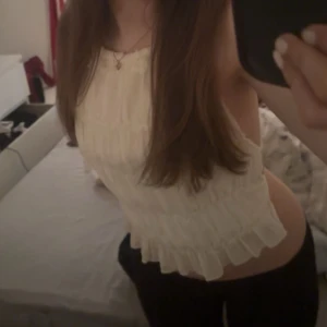 Vit volangig ärmlös blus - Säljer en söt vit blus med volangdetaljer och ärmlös design. Toppen har en rak halsringning och är lätt rynkad för en luftig känsla. Perfekt att styla med jeans eller kjol för en fräsch look.