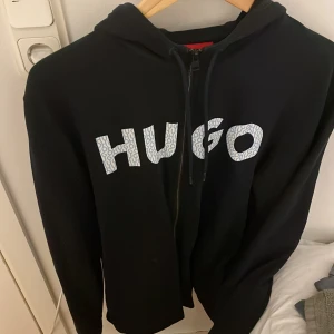 Svart hoodie med dragkedja från HUGO - Svart hoodie från HUGO med vit logga framtill i coolt mönster. Tröjan har huva med snörning, dragkedja hela vägen och långa ärmar. Perfekt för dig som gillar streetstyle och vill ha något bekvämt men ändå snyggt.
