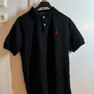 Svart pikétröja från Polo Ralph Lauren - Svart pikétröja från Polo Ralph Lauren med klassisk krage, två vita knappar och röd broderad logga på bröstet. Tillverkad i mjuk bomull och har korta ärmar. Perfekt för en clean och stilren look. Sprillans ny. Finns 2. En för 399 två för 599