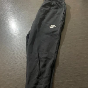 Svarta mjukisbyxor från Nike XS - Svarta mjukisbyxor från Nike i storlek XS med klassisk vit logga på vänster ben. Byxorna har resår i midjan med snörning och är tillverkade i mjukt material som passar perfekt för chill eller träning. Snygg och enkel design med fickor på sidorna.