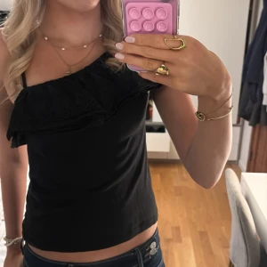 Svart oneshoulder topp från KappAhl - Svart oneshoulder topp från KappAhl med volangdetalj över bröstet och axeln. Toppen är ärmlös med smal axelrem på ena sidan och bred volang på andra. Super fin och perfekt till fester💕💕