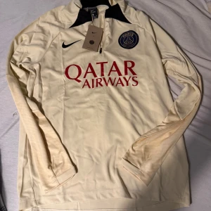 PSG långärmad fotbollströja Nike - Paris Saint-Germain långärmad fotbollströja i ljusbeige med svart krage och klubbmärke på bröstet. Röd Qatar Airways-logga framtill och Nike Swoosh. Tillverkad i lätt och ventilerande material, perfekt för träning eller match.
