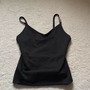 Svart linnetopp med strassband - Svart linnetopp med smala axelband dekorerade med strassdetaljer. Toppen har en enkel, stilren design med v-ringning och ärmlös passform. Perfekt för dig som gillar en clean look med lite extra bling på axelbanden.