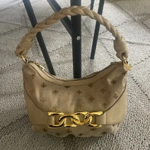 Beige handväska Friis & Company - Snygg beige handväska från Friis & Company med flätat handtag och guldiga detaljer. Väskan har ett diskret mönster, dragkedja upptill och en stor guldfärgad kedja framtill. Materialmix av textil och skinnimitation ger en lyxig känsla. Små defekter såsom sista bilden. Endast swish betalning skickas spårbart 59kr