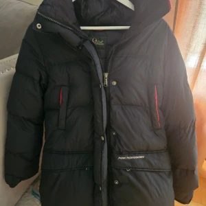 Svart dunjacka Peak Performance - Svart dunjacka från Peak Performance, modell Snowline J. Jackan har röda detaljer vid fickorna och på huvan, broderad logga på axeln och dragkedja framtill. Yttermaterial i polyamid och fyllning av 80% dun och 20% fjäder. Perfekt för kalla vinterdagar. Nypris 4800:-