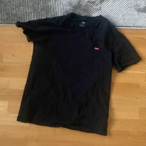 Svart t-shirt från Levi's - Svart t-shirt från Levi's med klassisk passform och liten röd logotyp på bröstet. Tillverkad i mjuk bomull och har rund halsringning samt korta ärmar. Perfekt basic-plagg med stilren look.