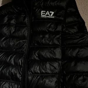 Svart dunväst från EA7 Emporio Armani - Snygg svart dunväst från EA7 Emporio Armani med quiltad design och logga på bröstet. Västen har dragkedja framtill och är tillverkad i polyester. Perfekt för dig som vill ha en clean och sportig look med premiumkänsla.