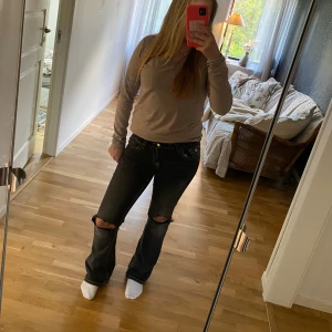 Svarta bootcut jeans med slitna knän - Säljer ett par svarta jeans med bootcut-modell och slitna hål på båda knäna. Jeansen har tydliga kontrastsömmar och klassiska bakfickor med lock och knapp. Materialet är jeans och passformen är normal. Perfekta för dig som gillar en avslappnad och trendig stil.