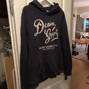Mörkblå hoodie från Ralph Lauren - Säljer en mörkblå hoodie från Ralph Lauren Denim & Supply med stor vit logga framtill. Tröjan har huva och en stor magficka, perfekt för en avslappnad och cool stil. Passar dig som gillar streetwear och klassiska märken.