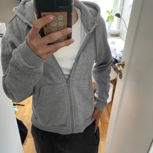 Grå hoodie från Adidas med dragkedja - Mysig grå hoodie från Adidas med dragkedja och huva. Tröjan har två stora fickor framtill och ribbade muddar vid ärmslut och nederkant. Insidan är fodrad med mjukt material som håller dig varm. Perfekt för chill dagar eller när du vill vara bekväm.
