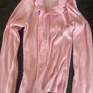 Rosa plisserad skjorta med knappar - Säljer en snygg rosa skjorta med plisserad struktur och klassisk krage. Skjortan har långa ärmar och stängs med vita knappar framtill. Materialet är glansigt och känns mjukt mot huden, perfekt för dig som vill sticka ut lite extra.