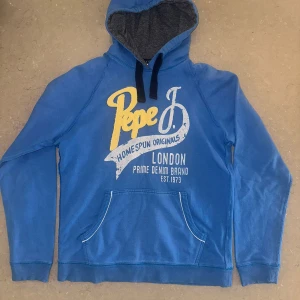 Blå hoodie från Pepe Jeans London - Snygg blå hoodie från Pepe Jeans London med tryck i gult och vitt framtill. Tröjan har huva med mörkblå snören, känguruficka och ribbade muddar. Slim fit-modell i mjuk bomull, perfekt för en avslappnad stil.