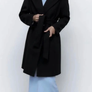 Svart kappa med bälte och knappar - Stilren svart kappa med klassisk krage, knytskärp i midjan och dubbla knappar framtill. Långärmad och rak modell som ger en clean och snygg siluett.  Är i bra skick. 