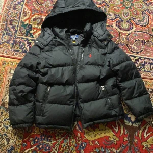 Svart pufferjacka Polo Ralph Lauren - Svart pufferjacka från Polo Ralph Lauren med huva och dragkedja. Jackan har en röd broderad logga på bröstet, två sidofickor med dragkedja och quiltad design. Perfekt för kalla dagar och riktigt snygg.