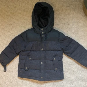 Svart pufferjacka från Quiksilver - Snygg svart pufferjacka från Quiksilver med huva och tryckknappar framtill. Jackan har två stora fickor med lock och är vadderad för extra värme. Perfekt för kalla dagar och har en stilren look med diskreta detaljer.