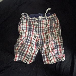 Rutiga shorts från Divided H&M - Snygga rutiga shorts från Divided by H&M i bomull. Färgerna är vit, blå och röd med klassiskt rutmönster. Shortsen har flera fickor och knappar framtill, samt en avslappnad passform. Perfekta för sommardagar och chill häng.