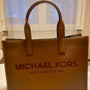 *NY*Cooper Michael Kors Luggage Structured Tote, Presenttips Bag, Presenttips  - Snygg och rymlig tote bag från Michael Kors i brunt skinn med präglad logga framtill. Ny oanvänd med prislapp. Unisex. Perfekt som weekendväska. Ett stort fack i mitten, ett mindre fack på sidan (passar laptop) och ett litet fack med blixtlås. Stängs med magnetlås. Ord pris: 6100 sek, Outlet: 4060 sek, Pris nu: 1500 sek.   Stay organized all day long with this generously sized carryall, featuring our embossed brand lettering. Crisp and minimalist in its design. 