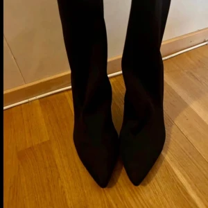 Svarta stövletter med klack Storlek 39 - Snygga svarta stövletter med hög klack och spetsig tå. Skorna har en stilren design och är tillverkade i ett material som ser ut som mocka. Perfekta för dig som vill ha en edgy och trendig look.