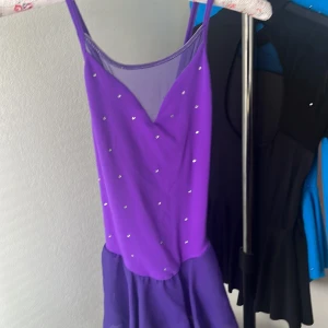 Lila konståkningsklänning med strassdetaljer - Superfin lila konståkningsklänning med glittrande strassdetaljer och mesh vid urringningen. Klänningen har smala axelband som korsas i ryggen och en flowy kjol i chiffongliknande material. Perfekt för dig som vill sticka ut på isen!