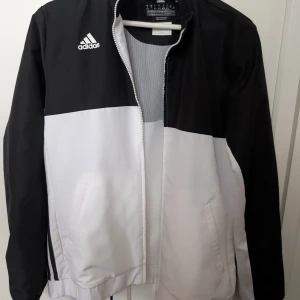 Svartvit vindjacka från Adidas - Snygg svartvit vindjacka från Adidas med klassisk logga på bröstet. Jackan har dragkedja framtill, två fickor och elastiska muddar. Tillverkad i polyester och har en sportig look med färgblock-design. Perfekt för dig som gillar streetwear och sportig stil. Storlek L