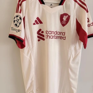 Liverpool Champions League tröja Isak 9 - Säljer en officiell Liverpool Champions League fotbollströja från Adidas med Isak och nummer 9 på ryggen. Tröjan är vit med röda detaljer, klubbmärke och sponsortryck framtill samt Champions League-märke på ärmen. Tillverkad i lätt och ventilerande material.