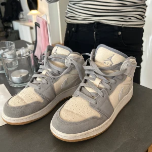 Nike Air Jordan 1 i grått och beige - Säljer ett par Nike Air Jordan 1 med snygg grå mocka och beige canvas. Skorna har klassisk snörning, gummisula i ljusbrunt och ikoniska Jordan-loggan på plösen. Perfekta för dig som gillar stilrena sneakers med retrovibe. Modellen heter Jordan 1 coconut milk
