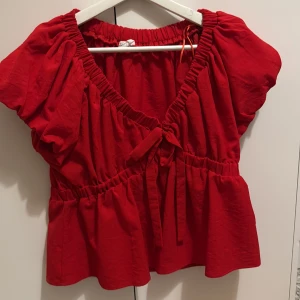 Röd blus med volang från Lushana Sky - Snygg röd blus från Lushana Sky med korta puffärmar, v-ringning och volangkant nertill. Toppen har resår vid halsringning och midja samt en söt knytning framtill. Perfekt för dig som vill ha en färgstark och luftig topp.