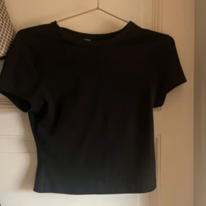 Svart croppad topp från SHEIN - En svart, enkel croppad topp från SHEIN i storlek XS. T-shirten har rund halsringning och korta ärmar. Perfekt att matcha med jeans eller kjol för en clean look. Gjord i mjukt material som känns skönt mot huden.