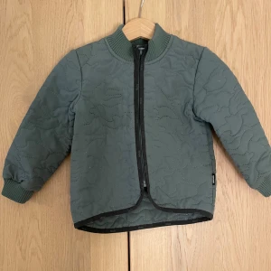 Quiltad grön jacka från Molo stl 86/92 - Quiltad grön jacka från Molo med svarta detaljer och dragkedja framtill. Jackan har ribbade muddar, rundad hals och en reflex stjärndetalj på ryggen. Perfekt för kyliga dagar och har en stilren look med snygg struktur.