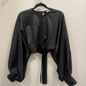 Svart croppad blus från NA-KD - Svart croppad blus från NA-KD i glansigt material med vida ballongärmar och knappar vid ärmslut. Toppen har öppen rygg med knytband.