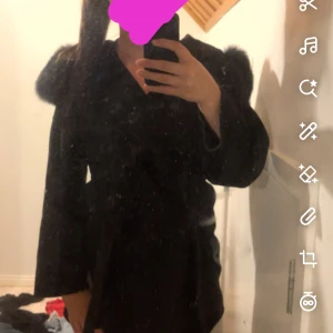 Svart pälsjacka med bälte - Säljer en svart pälsjacka med breda ärmar och markerade axlar. Jackan har ett bälte i midjan som ger en snygg siluett och är tillverkad i mjukt pälsmaterial. Perfekt för dig som vill sticka ut med en lyxig vibe.