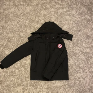 Canada goose Wyndham Jacka-svart-S - Säljer en svart dunjacka från Canada Goose i storlek S. Jackan har avtagbar huva med päls, klassisk logga på ärmen och flera fickor. Tillverkad i polyester och fodrad med dun och fjädrar. Perfekt för kalla vinterdagar och riktigt snygg streetstil.