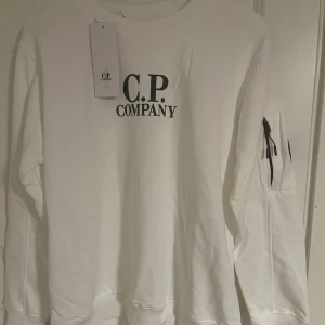 Vit sweatshirt från C.P. Company - Vit sweatshirt från C.P. Company med svart logga på bröstet och ikonisk linsdetalj på ärmen. Tröjan har rund hals, lång ärm och dragkedja på ärmfickan. Perfekt för dig som gillar streetwear och stilrena plagg. Den är M men passar även S