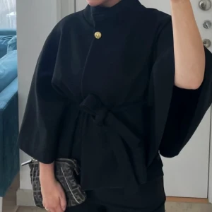 Svart poncho med knytband från Missay - Stilren svart kappa från Missay med vida ärmar, hög krage och ett snyggt knytband i midjan. Guldknapp vid halsen ger en exklusiv touch. Perfekt för dig som vill ha en trendig och elegant look. Använd knappats eftersom den har vart kort för mig inga defekter alls🩷