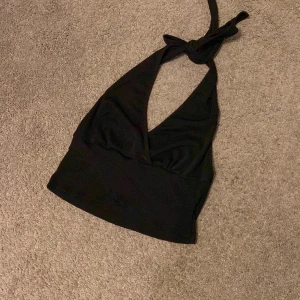 Svart halterneck linne med knyt - Säljer en svart halterneck linne med djup v-ringning och knyt i nacken. Toppen är croppad och har en bredare kant nedtill för en snygg passform. Perfekt för dig som gillar stilrena och enkla plagg med edge.