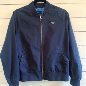 Marinblå Harrington-jacka från Gant - Snygg marinblå Harrington-jacka från Gant med klassisk ribbad krage, muddar och dragkedja framtill. Jackan har två snedställda fickor och diskret Gant-logga på bröstet. Insidan är silkeslent fodrad i ljusblått med stort GANT-tryck.  Tidlös, klassisk design som ger en uppklädd men avslappnad look. Storlek 158-164, passar även vuxen XS-S. Endast använd en gång. Nypris 1750kr.
