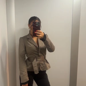 Beige kavaj med bälte och knappar - Snygg beige/grå kavaj med markerad midja tack vare bälte och dekorativa knappar framtill. Klassisk krage och långa ärmar, perfekt för en stilren look. Passformen är normal och jackan har en elegant siluett.