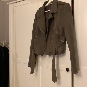 Beige mocka bikerjacka från NLY Trend - Säljer en snygg beige bikerjacka i mocka från NLY Trend, storlek 38. Jackan har klassisk krage, dragkedjor på fickorna, bälte i midjan och coola detaljer på axlarna. Perfekt för dig som vill ha en stilren och trendig look.