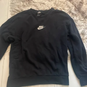 Svart Nike sweatshirt med logga - Svart sweatshirt från Nike med klassisk vit broderad logga på bröstet. Tröjan har rund halsringning och långa ärmar, perfekt för en sportig och avslappnad stil. Mjuk insida och ribbade muddar vid ärmslut och nederkant.