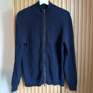 Massimo Dutti kofta  - Säljer en mörkblå stickad kofta med hel dragkedja framtill. Koftan har en snygg struktur i materialet, ribbade muddar och hög krage. Perfekt för lager-på-lager under kyliga dagar.