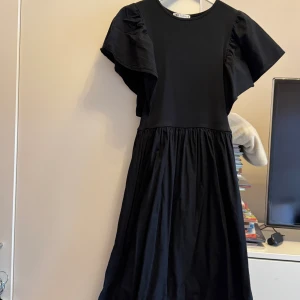 Svart klänning med volangärm från Zara - Svart klänning från Zara med rund halsringning och breda volangärmar. Modellen har en markerad midja och ett lätt utställt kjolparti. Tillverkad i mjuk bomull med lite stretch för extra komfort. Perfekt för dig som gillar en stilren och trendig look.