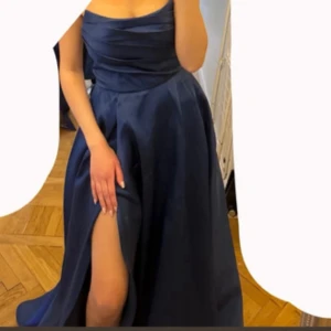 Marinblå långklänning med slits - Säljer en elegant marinblå långklänning med axelbandslös design och snygg slits framtill. Klänningen har ett glansigt tyg som ger en lyxig känsla och ett figurnära liv med draperade detaljer. Perfekt för dig som vill sticka ut på bal eller fest!
