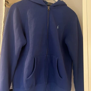 Blå hoodie från Polo Ralph Lauren - Säljer en blå hoodie från Polo Ralph Lauren med dragkedja framtill och klassisk huva. Tröjan har två stora fickor fram och den ikoniska lilla loggan broderad på bröstet. Perfekt för chill dagar eller när du vill ha en enkel och snygg look.