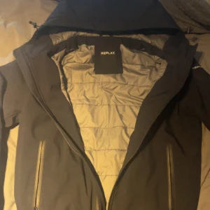 Svart pufferjacka från Replay - Snygg svart pufferjacka från Replay med huva och dragkedja framtill. Jackan har två praktiska fickor med dragkedja och quiltad insida i ljusgrått. Perfekt för kyliga dagar och har en stilren look som passar till det mesta.