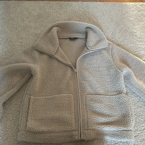 Beige teddyjacka från 157, XS - Mysig beige teddyjacka från 157 med dragkedja framtill och två stora fickor. Jackan har hög krage och är i fluffigt teddy-material som håller dig varm. Perfekt oversized look och passar till jeans eller mjukisbyxor.