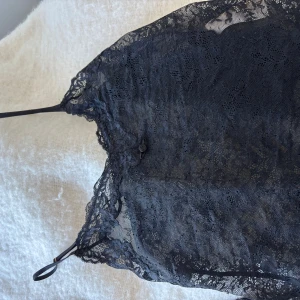 Svart spetsig linnetopp med tunna band - Säljer en svart linnetopp i genombruten spets med tunna axelband och v-ringad hals. Toppen har ett feminint blommigt spetsmönster och är lätt transparent. Perfekt för dig som gillar en chic och edgy stil.