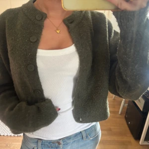 Olivgrön kofta från Zara i ullmix - Säljer en olivgrön kofta från Zara i ullmix. Koftan är croppad med stora knappar framtill, ribbade muddar och rund halsringning. Perfekt att slänga över ett linne eller t-shirt för en chill look. Den har lite noppror 