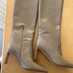 Beige croc boots från NA-KD, stl 41 - Snygga beige boots från NA-KD i storlek 41 med croc-mönstrad yta. Modellen har spetsig tå, hög klack och dragkedja på insidan. Tillverkade i syntetmaterial med en lyxig look. Perfekta för dig som vill sticka ut med trendiga stövlar. Väldigt lite skav på insidan upptill se bilder. Väldigt lite använda